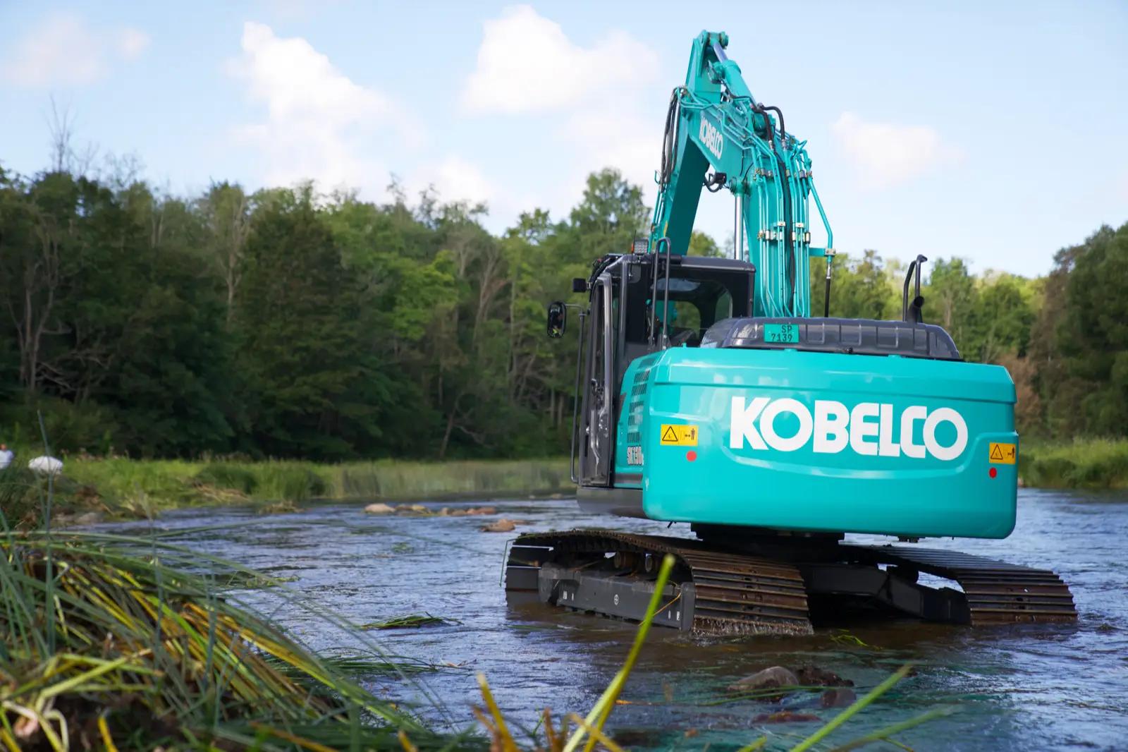 Kobelco Kettenbagger der SK130-Baureihe steht in einem flachen Fluss