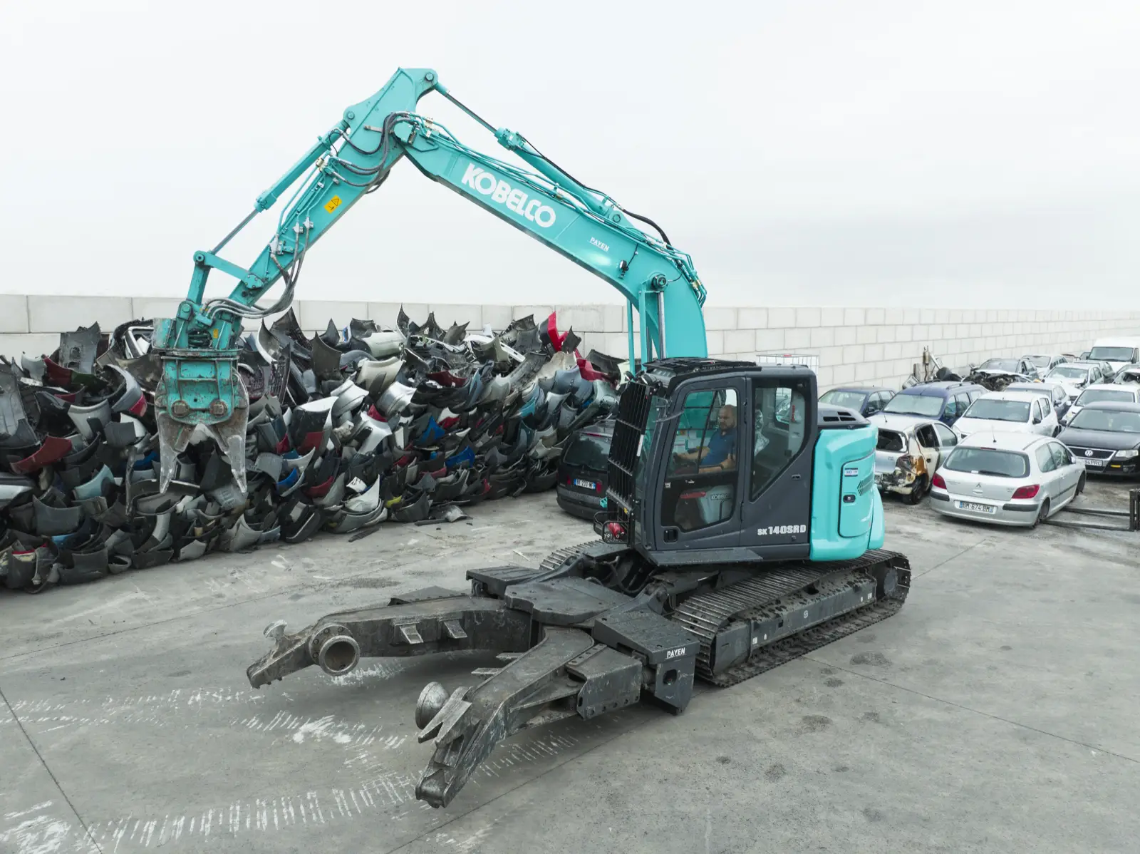 Kobelco SK140SRD mit Klemmarm vor Fahrzeugteilen