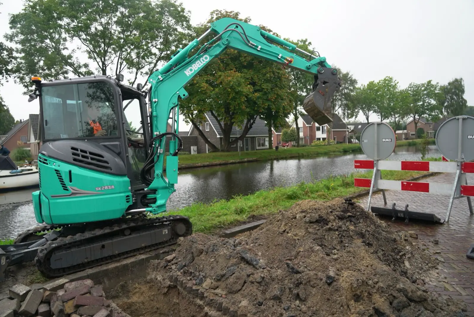 Kobelco SK26SR mit Kabine hebt an einem Kanal einen Graben neben der Straße aus